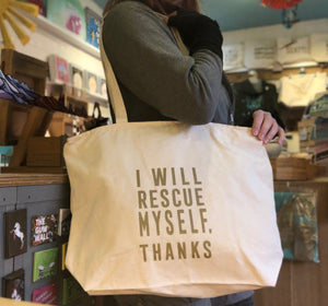 A person modeling a cream colored canvas tote bag that says "I will rescue myself, thanks" in gold lettering.