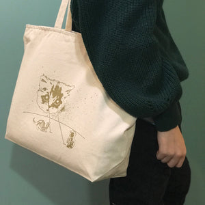 A person carrying a beige cotton tote bag with a gold screen printed cat design on one side.