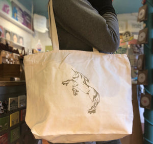 A cream colored tote bag with a roller skating unicorn printed in gold ink.