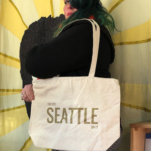 A person holding a tote bag that says "This says Seattle on it" in gold ink.