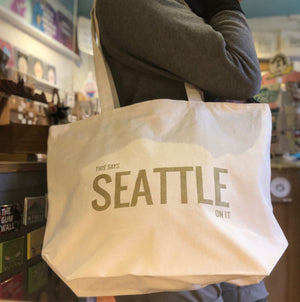 A person holding a cream colored tote bag that says "This says Seattle on it" in gold ink.