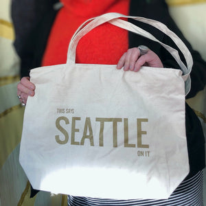 A person holding a cream colored tote bag that says "This says Seattle on it" with gold ink.
