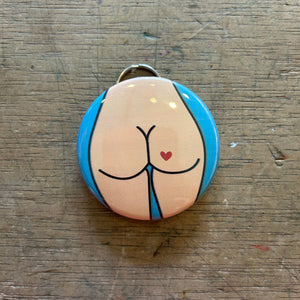Bottle opener keychain featuring a design of a light colored butt with a red heart tattoo on a blue background on top of a wooden surface.