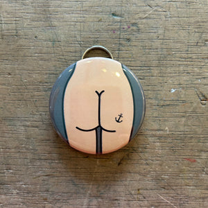 Bottle opener keychain featuring design of a light colored butt with an anchor tattoo on a gray background on top of a wooden surface.