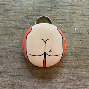 Bottle opener keychain featuring a design of a light colored butt with an anchor tattoo on an orange background on top of a wooden surface.