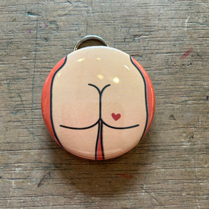Bottle opener keychain featuring a light colored butt with a red heart tattoo on an orange background on top of a wooden surface.