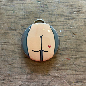 Bottle opener keychain featuring a design of a light colored butt with a red heart tattoo on a gray background on top of a wooden surface.