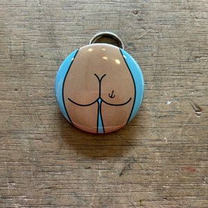 Bottle opener keychain featuring a design of a tan butt with an anchor tattoo on a blue background on top of a wooden surface.