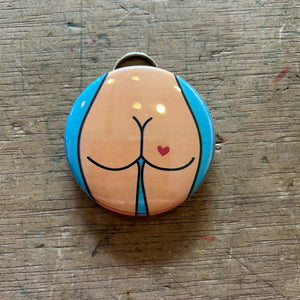 Bottle opener keychain featuring a design of a tan butt with a red heart tattoo against a blue background on top of a wooden surface.