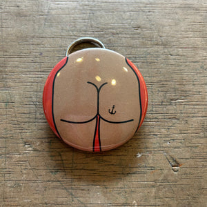 Bottle opener keychain featuring a design of a tan butt with an anchor tattoo against an orange background on top of a wooden surface.