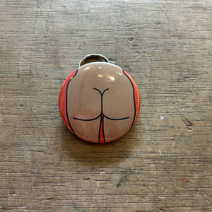 Bottle opener keychain featuring a design of a tan butt on an orange background on top of a wooden surface.