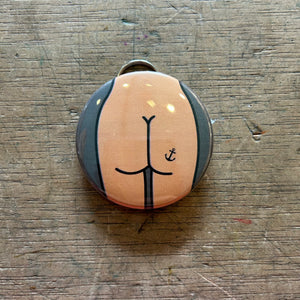 Bottle opener keychain featuring a tan butt with an anchor tattoo against a gray background on top of a wooden surface.