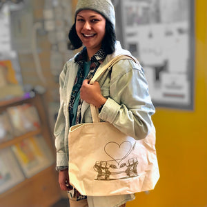 A smiling dark haired woman wearing a gray beanie is holding an off white color tote bag with two AT-ATs falling in love.