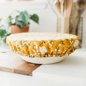 A yellow fabric stretch cover with a white floral pattern is stretched over a white bowl on a kitchen counter, with a wooden cutting board in the foreground and a softly blurred kitchen background.