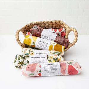 Several reusable bowl covers with printed fruit and floral designs are arranged in and around a woven wicker basket on a white surface against a white tiled wall.