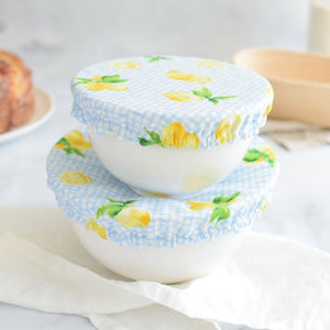 Two white ceramic bowls stacked on top of each other, covered with light blue and white gingham fabric lids printed with yellow lemons and green leaves, set on a white cloth in a softly blurred kitchen setting.