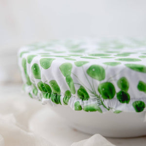 A close-up of a white fabric cover with a green leaf pattern stretched over a round container, set against a soft, light background.