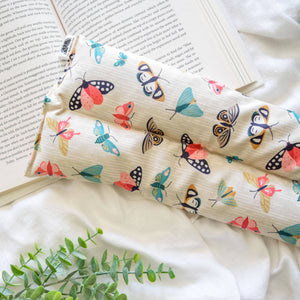 A hot/cold pack with a colorful moth print on a cream background, styled on top of an open book and white fabric with a green plant sprig.