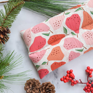 A hot/cold pack with a pear pattern in shades of pink, coral, and orange on a cream linen background, styled on a white marble surface with pine branches, pinecones, and red berries.