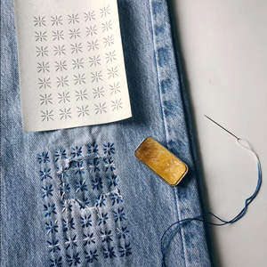 Stitched up example of mending transfers on a pair of jeans with a needle, thread, and tin of thread gloss.