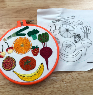 Stitched up embroidery designs of various fruits and vegetables in an orange embroidery hoop on a wooden background.