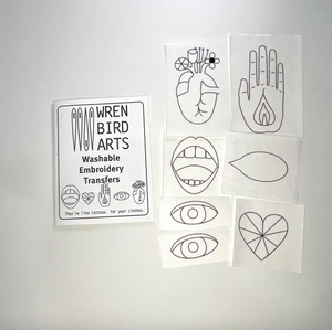 A set of white washable embroidery transfer sheets from Wren Bird Arts, featuring black line-art designs including a heart, hand, lips, eyes, and a heart motif, displayed next to a product label on a light gray background.