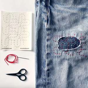 A stitched up example of one of the mending patterns on a pair of jeans with a needle and thread and a pair of scissors.