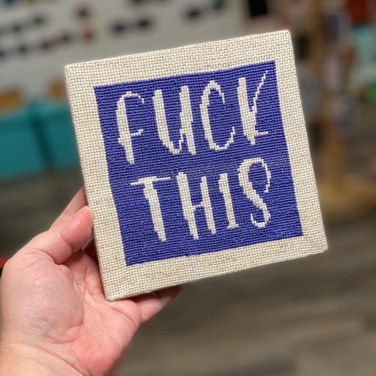 A cross stitch kit that says "Fuck This" Stitched in dark purple on an off-white canvas. 