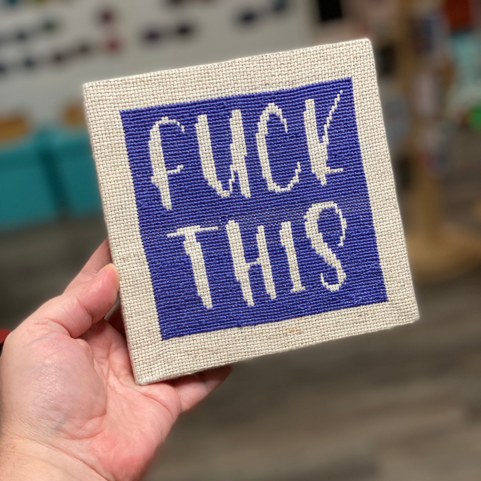 A cross stitch kit that says "Fuck This" Stitched in dark purple on an off-white canvas. 