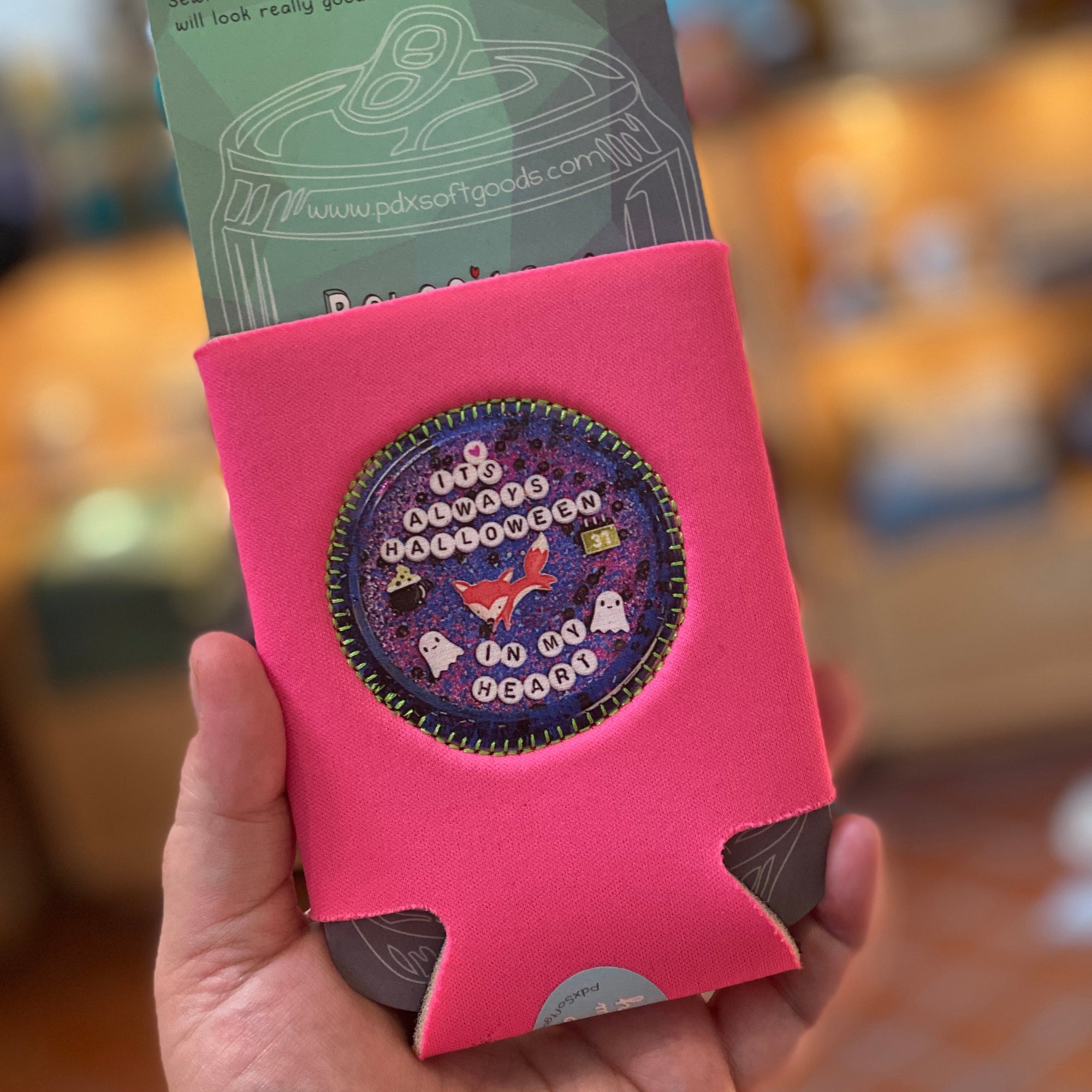 A hand is holding up a bright pink drink holder that says "It's  Always Halloween in My Heart"