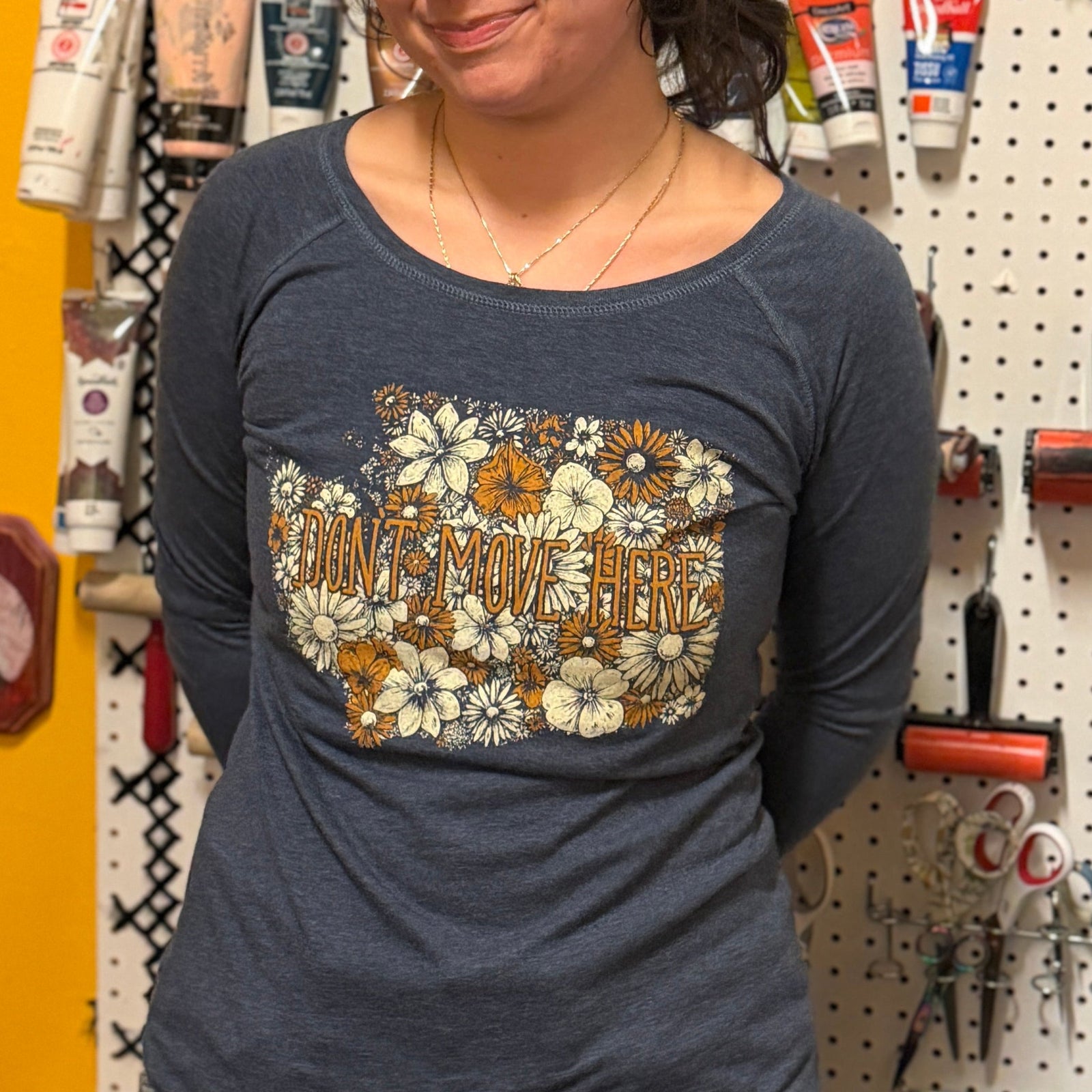 Woman wearing a blue shirt with floral design standing in front of a pegboard wall with tools and supplies.
