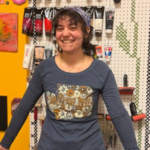 Person wearing a blue long-sleeve shirt with floral design and text, standing in front of a pegboard wall with various items.