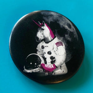 Against a bright blue background is a black and white unicorn with bright pink horn, mane and roller skates. He is holding an astronaut helmet and is wearing a space suit. It says his name is Moondancer. 