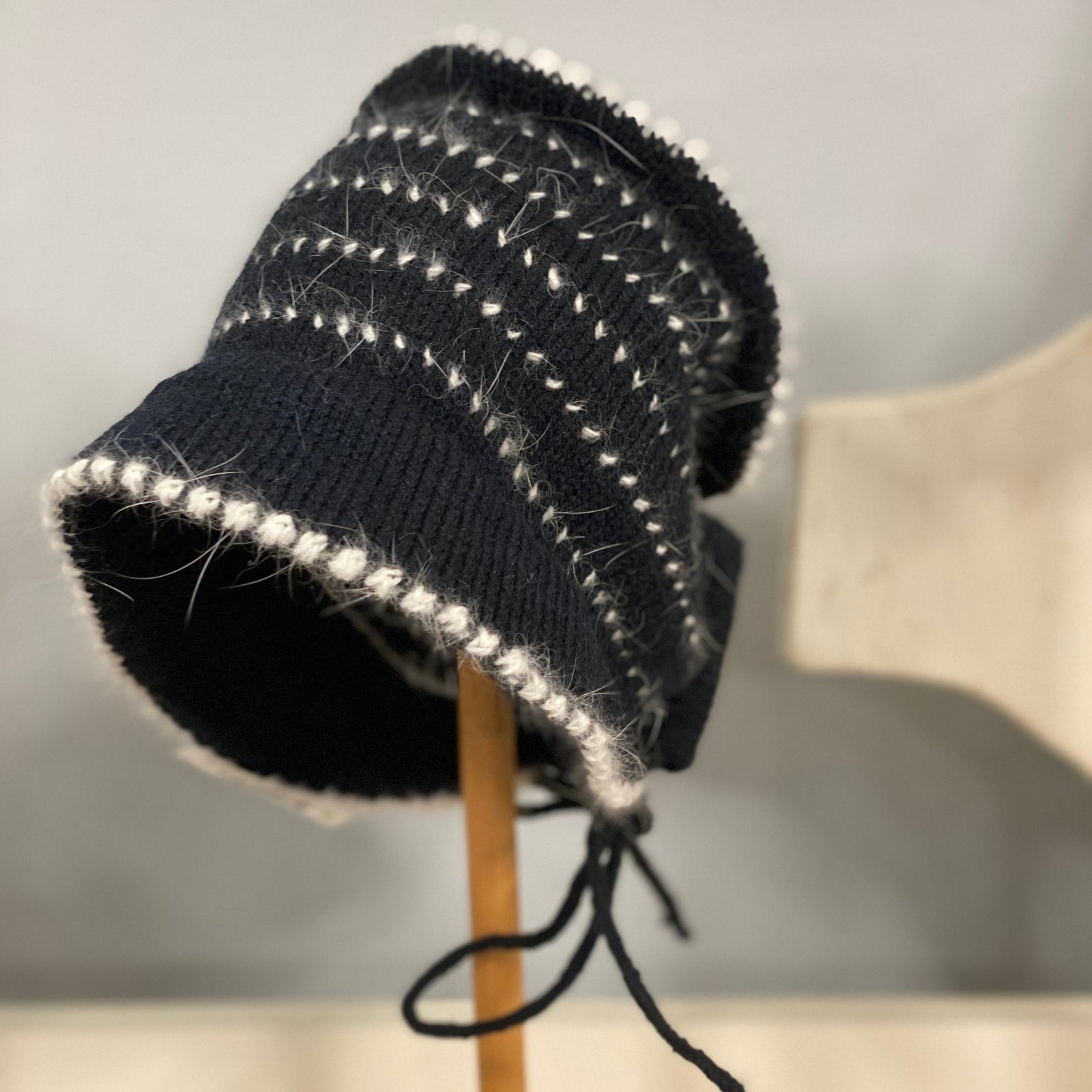 Black knitted hat with white trim on a wooden stand against a neutral background. 
