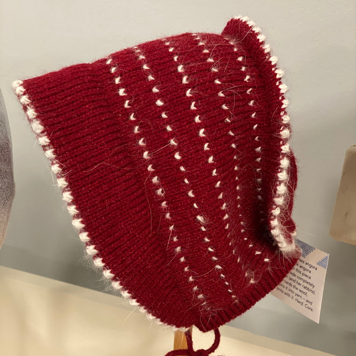 Red knitted bonnet with white trim on a wooden stand against a neutral background. 