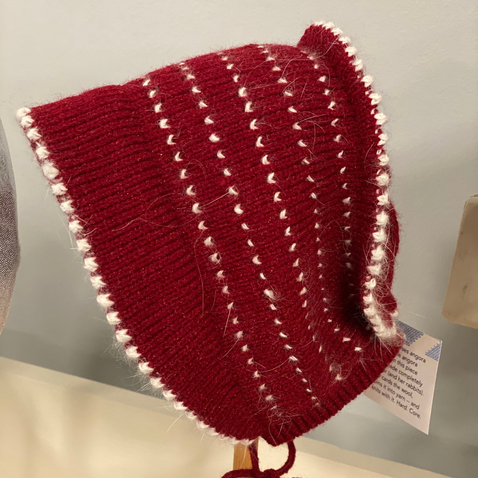 Red knitted bonnet with white trim on a wooden stand against a neutral background. 