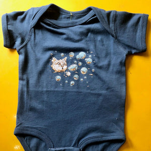 Baby bodysuit laid flat on a yellow table. The bodysuit has Ugly Baby's Bubble Cat design screenprinted on it. There is a large cloud cat blowing bubbles and inside the bubbles are tiny kittens. 
