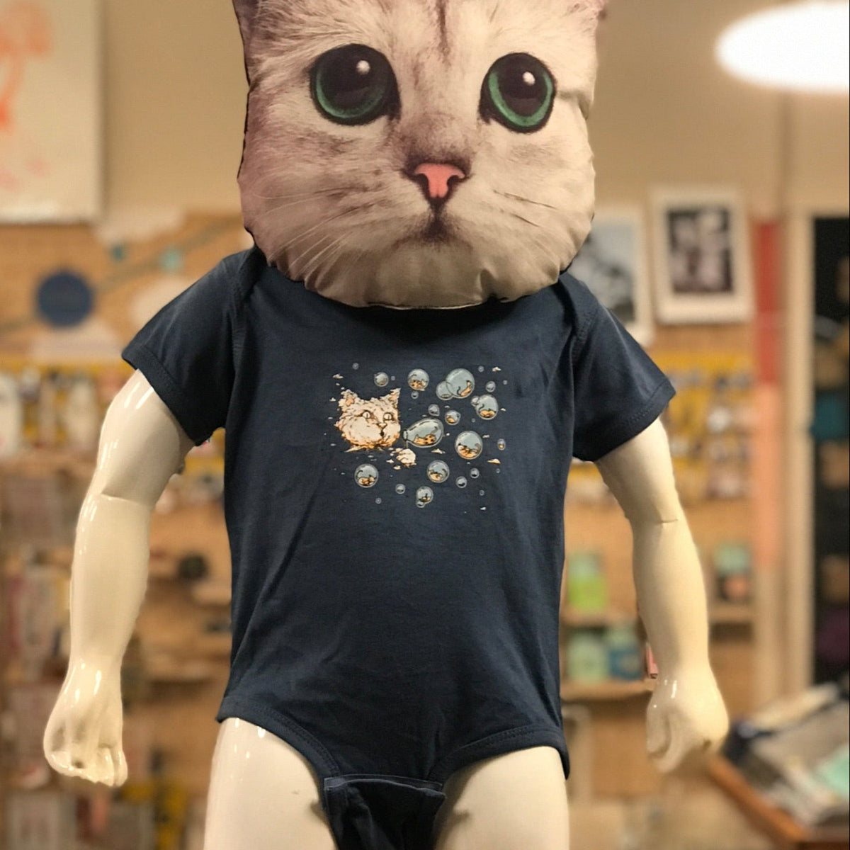 Mannequin with a cat head wearing a dark blue baby romper with Ugly Baby's Bubble Cat design in an indoor setting.