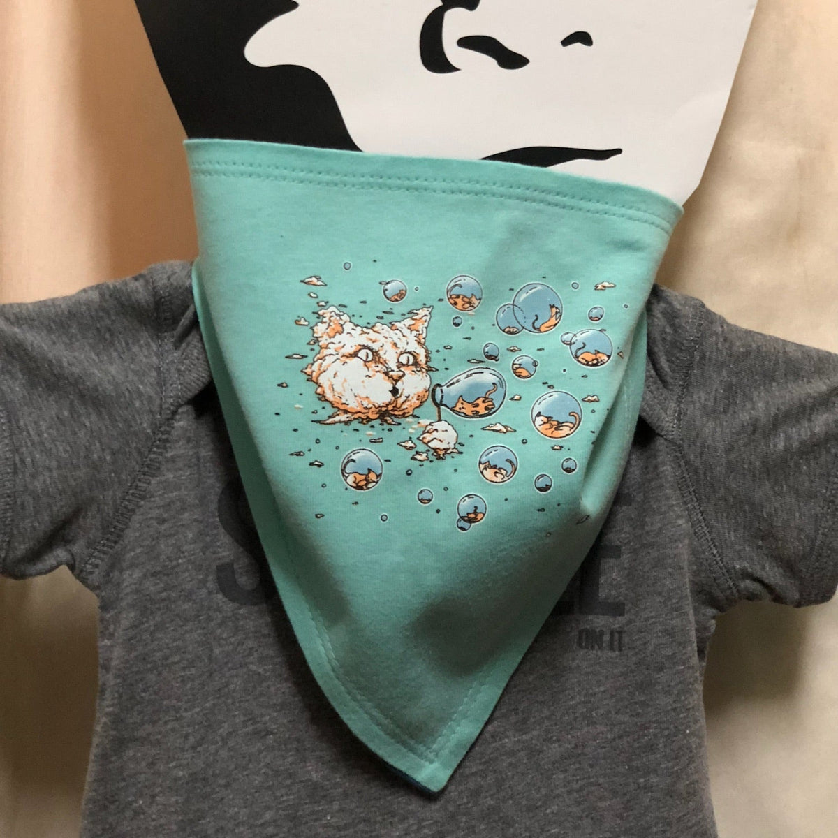 A mint colored bandana bib with our Bubble Cat design on it. A large cat-head-shaped cloud is blowing bubbles. Inside those bubbles are tiny kittens. 