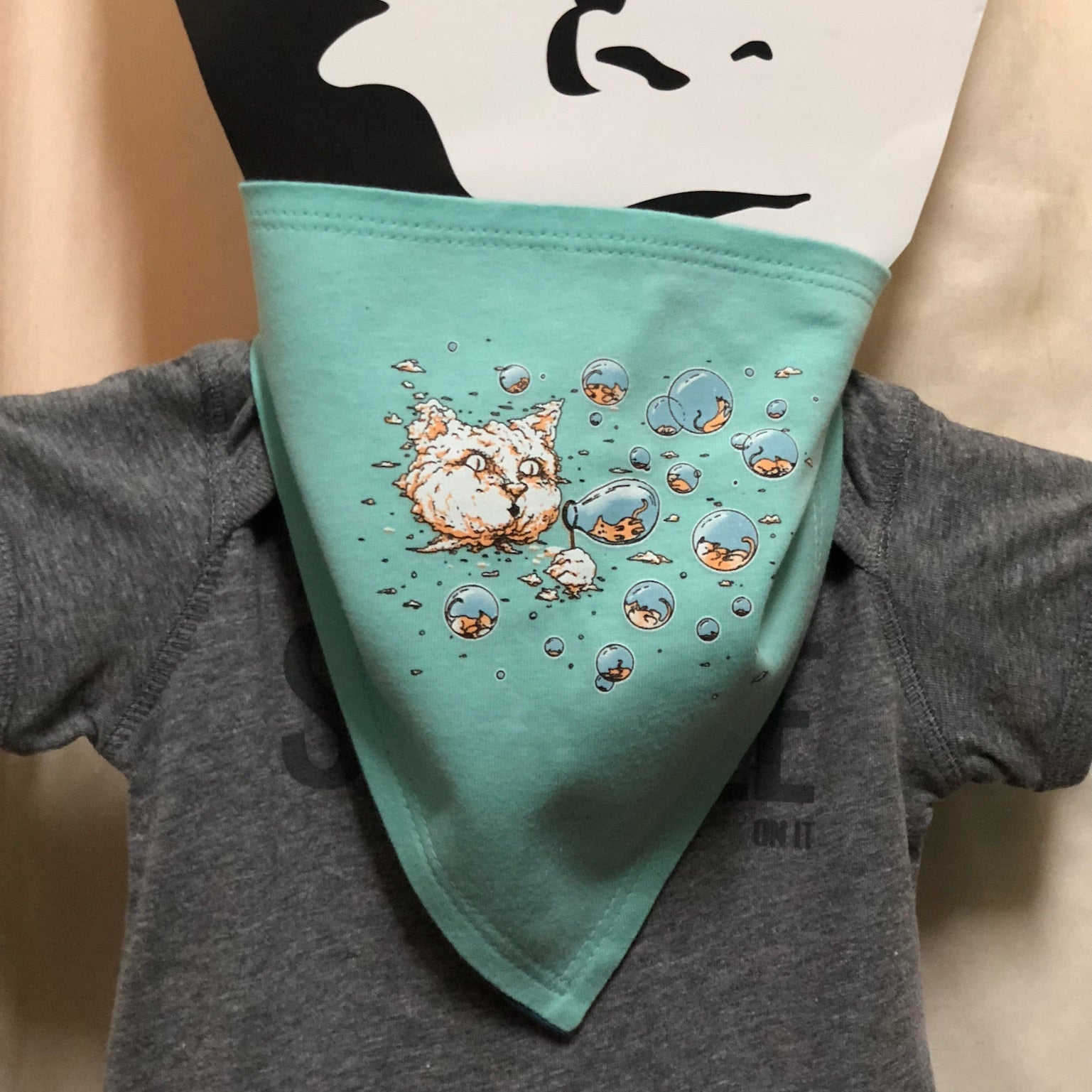 A mint colored bandana bib with our Bubble Cat design on it. A large cat-head-shaped cloud is blowing bubbles. Inside those bubbles are tiny kittens. 