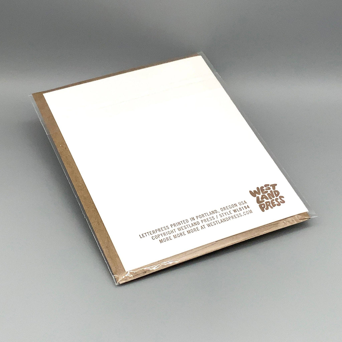 The back of a white greeting card with a kraft brown border, wrapped in clear plastic, photographed on a smooth gray studio background. The lower right corner shows the West Land Press logo and small printed text.