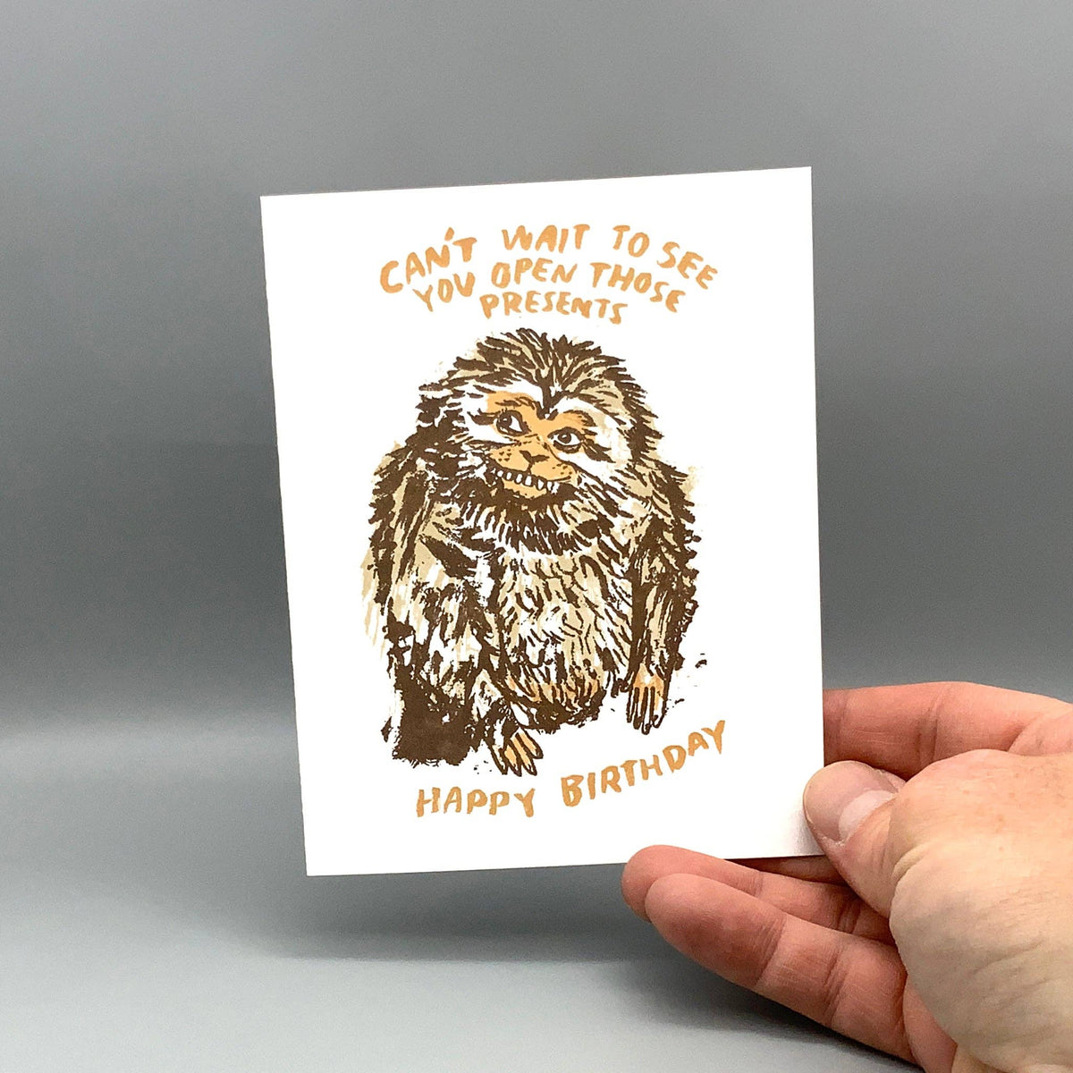 A hand holding a white greeting card with a brown sketch of a monkey and orange text reading 'CAN'T WAIT TO SEE YOU OPEN THOSE PRESENTS' and 'HAPPY BIRTHDAY' against a gray studio background.