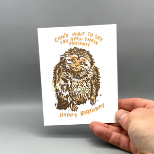A hand holding a white greeting card with a brown sketch of a monkey and orange text reading 'CAN'T WAIT TO SEE YOU OPEN THOSE PRESENTS' and 'HAPPY BIRTHDAY' against a gray studio background.
