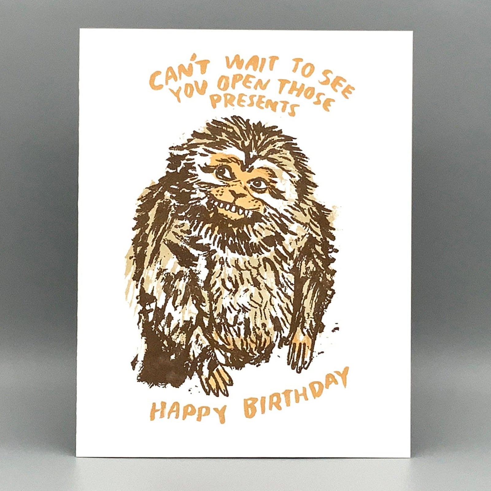 A white greeting card featuring a brown sketch-style illustration of a furry creature with the text 'CAN'T WAIT TO SEE YOU OPEN THOSE PRESENTS' above and 'HAPPY BIRTHDAY' below, photographed against a gray gradient background.