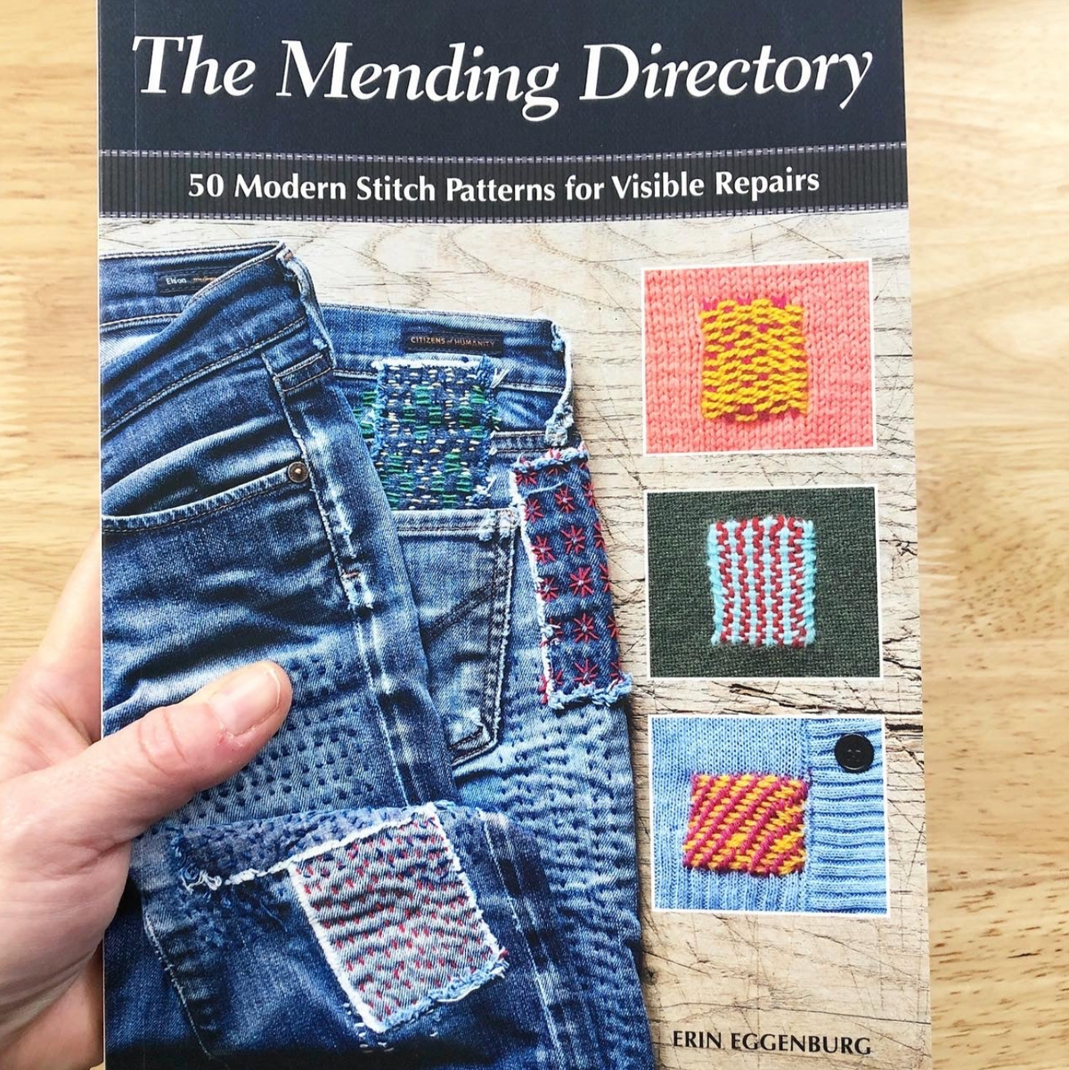 Cover of the book: The Mending Directory. It shows a hand holding up the book. The cover of the book has a few different examples of stitches on knitted items as well as denim.