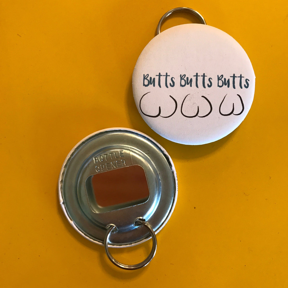 Metal bottle opener with a round keychain attached on an orange background. On the front it has three butts and says "Butts Butts Butts"