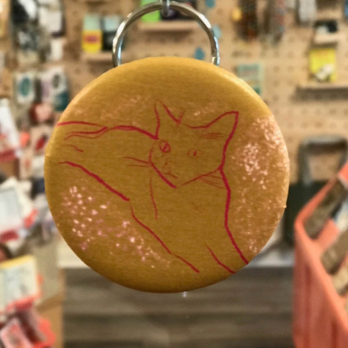 Round keychain with a red cat illustration on a yellow background, hanging in a store.