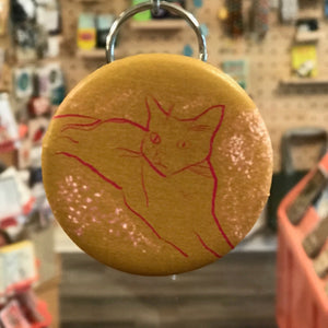 Round keychain with a red cat illustration on a yellow background, hanging in a store.