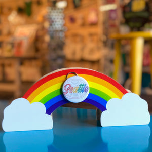 Rainbow-shaped object with 'Seattle' bottle opener keychain on a reflective surface, blurred store interior background. 