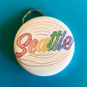 White bottle opener keychain that has Seattle written in rainbow colors & the letters are made out of raindrops. 